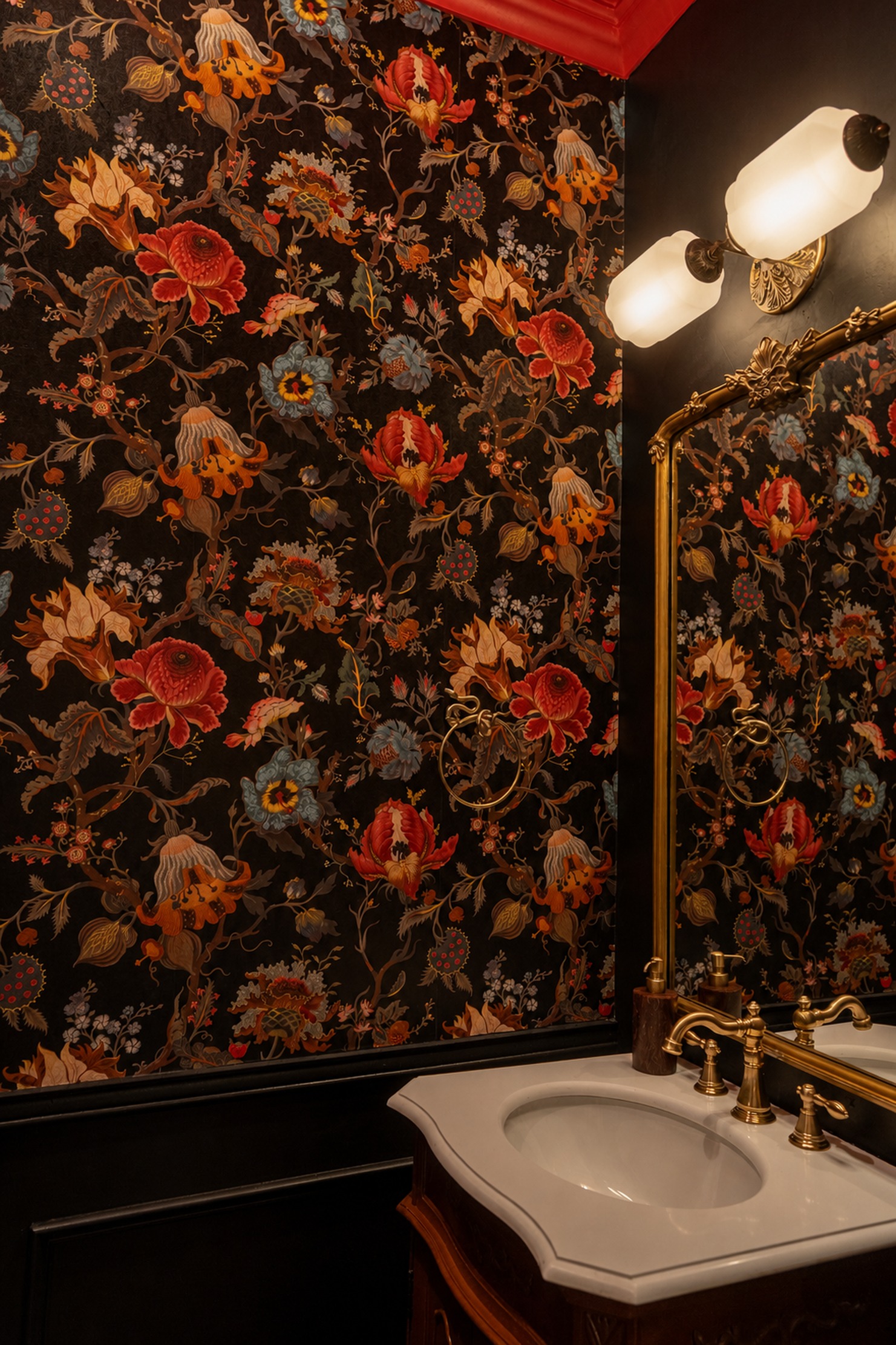 The powder room, dark floral wallpaper, brass sconce and faucet, a glimpse of red ceiling trim