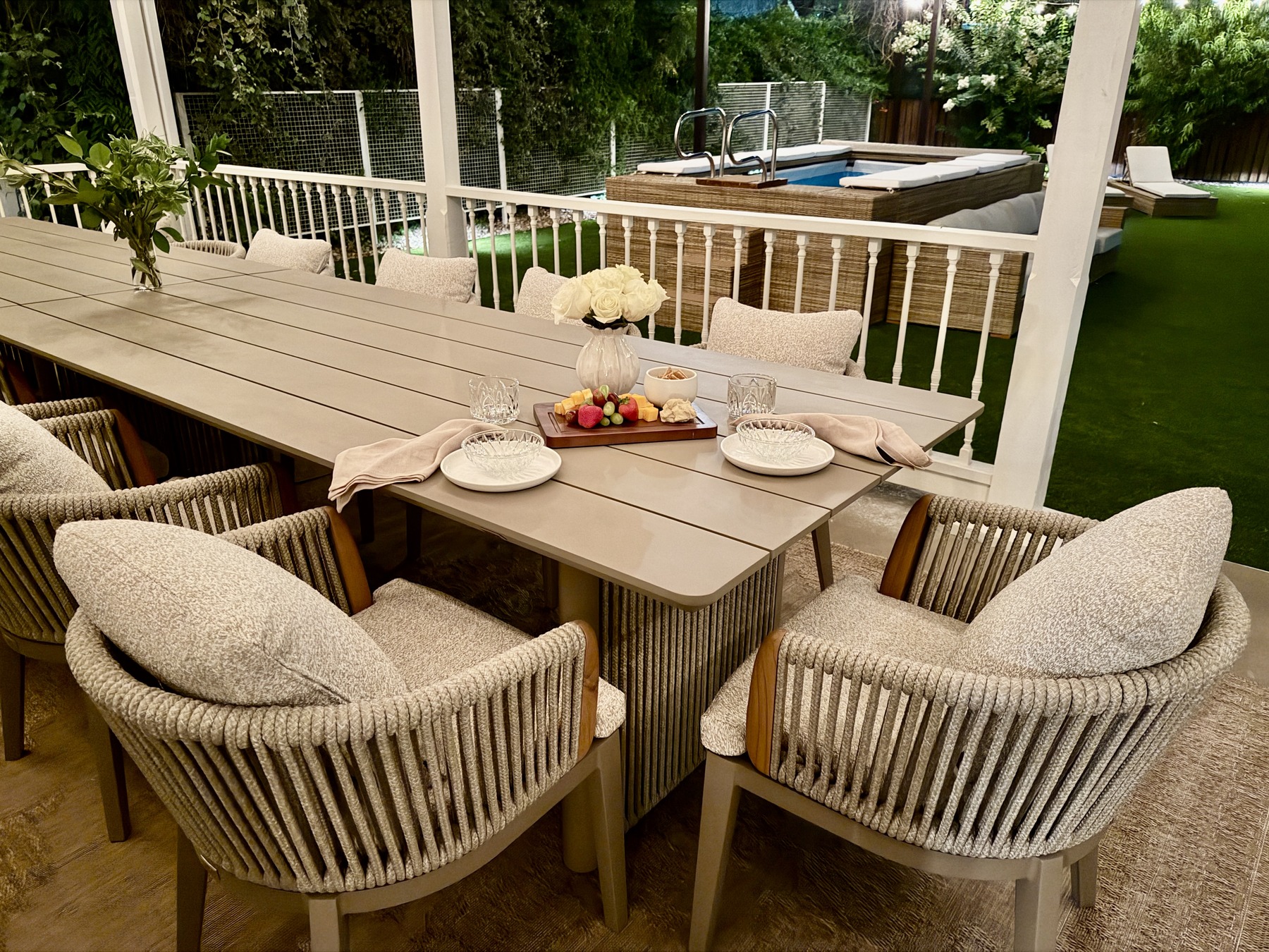 The covered dining table set for a group, roses and a shared board at the center, the pool and lounge seating visible beyond in daylight