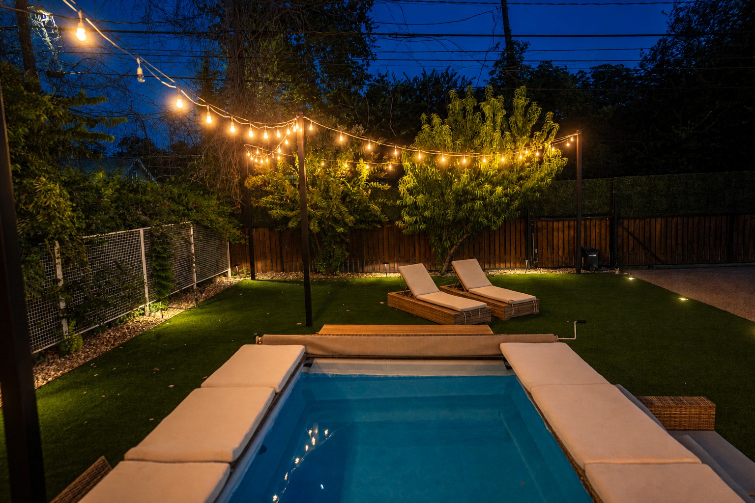 The pool at night, straight-on, string lights and trees glowing above the water