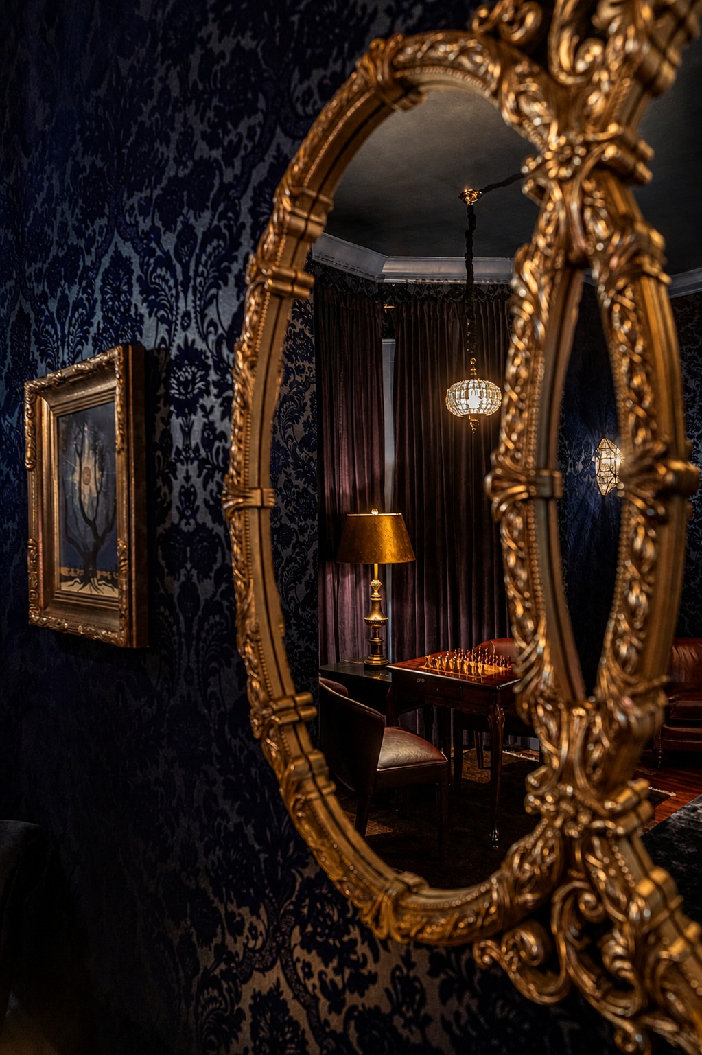 A closer oval mirror reflecting the chess table, lamp, and chandelier, a framed painting soft-focus in the foreground