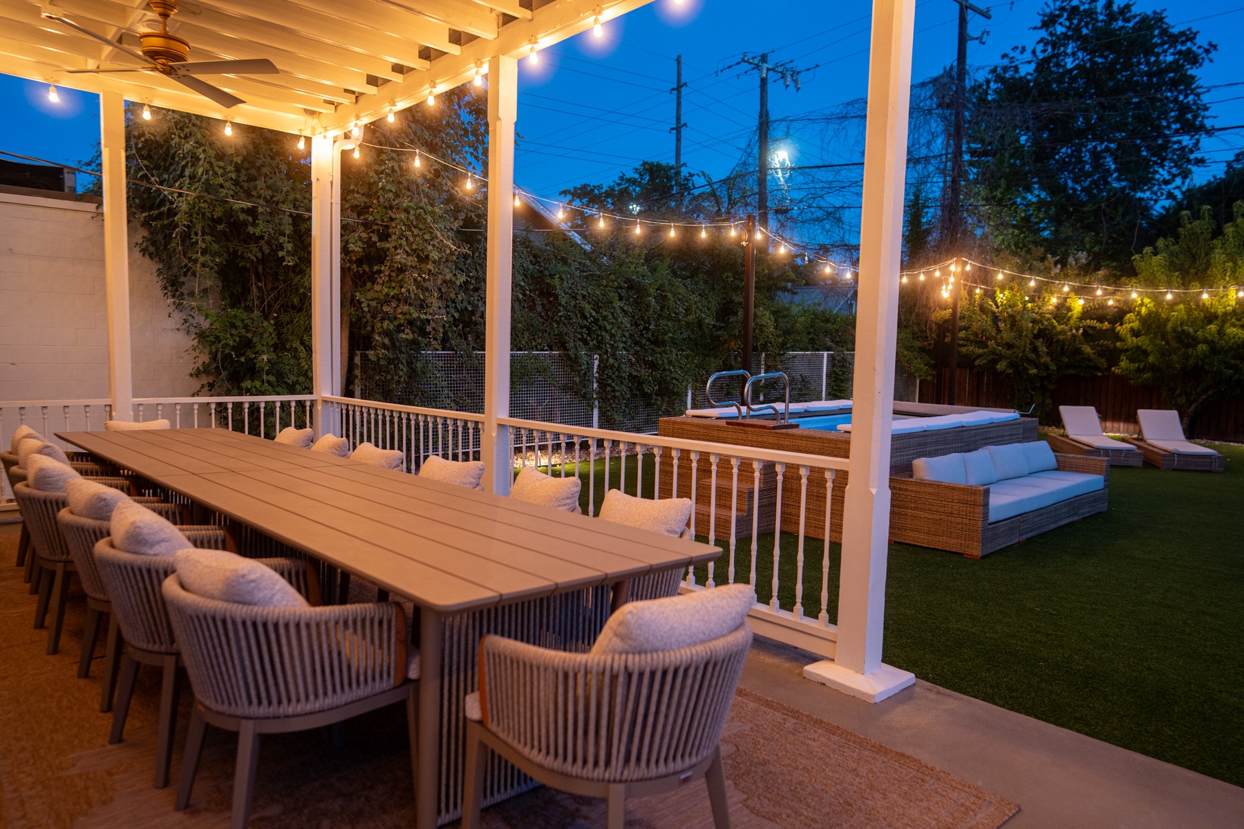 Under the pergola at dusk, the long dining table set for fourteen, string lights leading out to the pool and garden